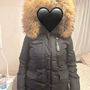 Svart dunjacka med päls från Hollies - Säljer en svart dunjacka från Hollies med stor, lyxig pälskantad huva. Jackan har flera fickor med lock och dragkedja samt markerad midja. Perfekt för kalla dagar och riktigt snygg siluett.