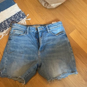 Blå jeansshorts från Bik Bok - Snygga blå jeansshorts från Bik Bok med råa, fransiga benslut och klassisk femficksmodell. Shortsen har en avslappnad passform och är perfekta för sommardagar. Tillverkade i slitstarkt denim med dragkedja och knapp framtill.