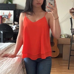 Skarpt orange linnetopp från Pieces - Säljer en snygg skarpt orange linnetopp från Pieces med tunna axelband och v-ringning. Toppen har dubbla lager tyg som ger en flowy och luftig känsla. Perfekt till jeans eller kjol för en fräsch look. Enkel och stilren design som är lätt att matcha.