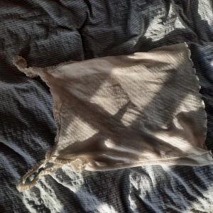 Ljusbeige ribbad linnetopp med spets - Supersöt ljusbeige linnetopp med ribbad struktur och tunna axelband dekorerade med spets. Toppen har vågiga kanter och en mjuk, stretchig känsla som gör den perfekt till jeans eller kjol.