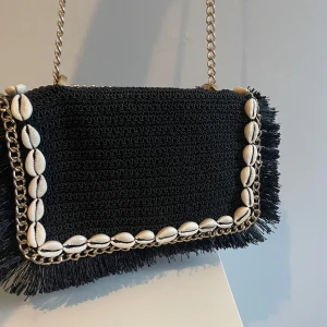 Svart axelväska med snäckskal och kedja - Säljer en svart axelväska med flätad struktur och fransar längs kanterna. Väskan har guldfärgad kedjerem och är dekorerad med snäckskal runt locket för en riktigt somrig vibe. Perfekt att slänga över axeln när du vill sticka ut lite extra.