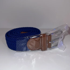 Blått flätat bälte  - Snyggt blått flätat bälte med bruna skinnliknande detaljer vid spännet och silverfärgat metallspänne. Perfekt accessoar för att lyfta din outfit med en färgklick.