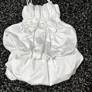 Vit kort klänning med rosett - Supersöt vit kort klänning i glansigt satinliknande material med stor rosett framtill. Axelbandslös modell med puffig kjol och draperad effekt. Perfekt för dig som vill sticka ut med en feminin och lekfull look.