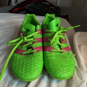 Gröna och rosa Adidas fotbollsskor - Säljer ett par riktigt coola Adidas fotbollsskor i neon-grönt med rosa ränder och snörning. Skorna har klassisk rund tå och är tillverkade i syntetmaterial med struktur för extra grepp på planen. Perfekta för dig som vill sticka ut på fotbollen!