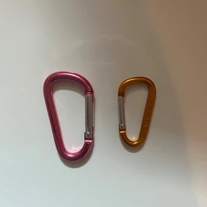Två karbinhakar i rosa och orange - Säljer två snygga karbinhakar i metall, en i metallic rosa och en i metallic orange. Perfekta att hänga på väskan för att fästa nycklar eller accessoarer. Smidiga och lätta att använda med fjäderlås i silver.