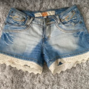 Jeansshorts med spets från Miss Chic - Snygga ljusblå lågmidjade jeansshorts från Miss Chic med broderade detaljer på fickorna och vit spetskant nertill. Shortsen har klassisk femficksmodell, gylf med knapp och bälteshällor. Perfekta för sommaren och ger en söt och trendig look.