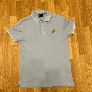 Ljusblå pikétröja från Lyle & Scott - Snygg ljusblå pikétröja från Lyle & Scott med klassisk krage, vita detaljer på krage och ärmslut samt den ikoniska gula örnloggan på bröstet. Tröjan har korta ärmar och är tillverkad i mjuk bomull som känns skön mot huden.