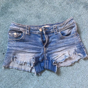 Blå jeansshorts med fransig kant - Snygga blå jeansshorts med fransig och sliten kant. Klassisk femficksmodell med knapp och dragkedja framtill. Perfekta för varma dagar och ger en cool, avslappnad vibe. Passar dig som gillar en lite tuffare stil.