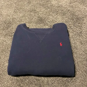 Mörkblå sweatshirt från Polo Ralph Lauren - Mörkblå sweatshirt från Polo Ralph Lauren med klassisk röd broderad logga på bröstet. Tröjan har rund halsringning, långa ärmar och ribbade muddar. Tillverkad i mjuk bomull som är skön att ha på sig. Perfekt för en chill och stilren look. Stl L barn 