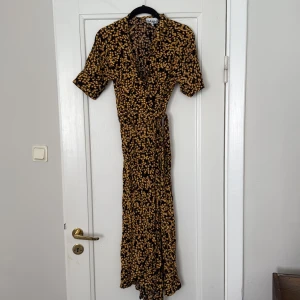 Ganni Svart långklänning med blommigt mönster  - Säljer en svart långklänning från Ganni med korta ärmar och blommigt mönster i gul. Omlottklänning. Storlek 40 men har passat mig som är en S/36