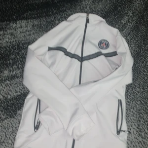 Vit psg Nike tech  - Snygg vit PSG vindjacka med svarta detaljer och dragkedjor. Jackan har klubbens emblem på bröstet, svarta ränder över bröstet och svarta dragkedjor på fickorna. Perfekt för dig som vill ha sportig stil och visa vilket lag du hejar på.Storlek 164 12-14.kommer ren