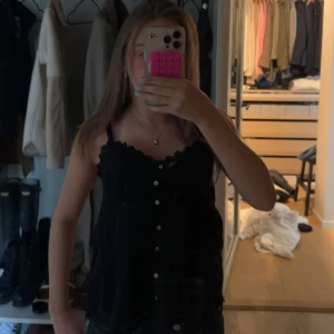 Svart linnetopp med spets och knappar - Säljer en svart linnetopp med smala axelband, dekorativa spetsdetaljer längs urringningen och knappar framtill. Toppen har en lätt volangig nederkant. Den är gammal från Zara och är i strl XS. 