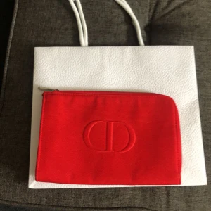 Röd Dior kuvertväska med dragkedja - Säljer en snygg röd kuvertväska från Dior i canvas med silverfärgad dragkedja och präglad CD-logga på framsidan. Perfekt storlek för det viktigaste och enkel att bära med sig. Insidan är helröd och väskan har en stilren look.