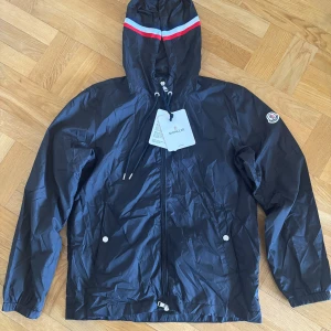 Moncler svart vindjacka med huva - Snygg svart vindjacka från Moncler med huva och dragsko. Jackan har dragkedja framtill, två fickor med tryckknappar och Moncler-logga på ärmen. Huvan har en röd, vit och blå rand som ger en sportig vibe. Perfekt för blåsiga dagar.