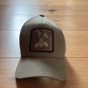 Beige keps med Wile E. Coyote-märke - Säljer en beige keps med mesh baktill och justerbar snapback. Framtill finns ett broderat märke med Wile E. Coyote. Perfekt för dig som gillar tecknade klassiker och vill ha en stilren accessoar.