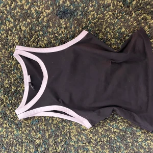 Svart croppad linnetopp med vita kanter - Snygg svart croppad linnetopp med vita kontrastkanter runt hals och ärmhål. Perfekt för sommaren och enkel att matcha med olika outfits. Modellen är ärmlös och har en sportig känsla.