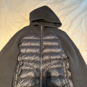 Svart black label Moncler hybridjacka med huva - Säljer en grå hybridjacka från Moncler med stickade ärmar och huva samt quiltad, dunfylld front. Jackan har dragkedja framtill och två fickor. Perfekt för dig som vill ha en snygg och sportig look med exklusiv känsla. Det finns kod som kan bekräfta att den är äkta skriv till mig så skickar jag det.