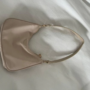 Beige axelväska i satinlook - Snygg beige axelväska med stilren design och guldig dragkedja. Väskan har en mjuk satinliknande yta och justerbart axelband. Extra axelrem medföljer för mer variation. Perfekt storlek för mobil, plånbok och andra essentials.