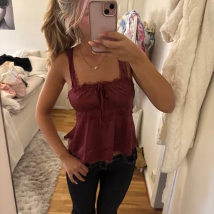 Vinröd linnetopp med spets och knyt - Supersöt vinröd linnetopp med spetsdetaljer och knyt framtill. Toppen har breda axelband, volangkant nertill och öppen rygg som ger en riktigt snygg look. Perfekt att styla med jeans eller kjol för en trendig vibe.