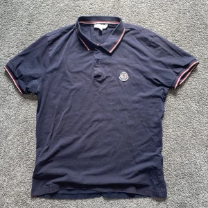 Mörkblå pikétröja från Moncler - Säljer en mörkblå pikétröja från Moncler med klassisk krage och knäppning framtill. Tröjan har röda och vita ränder på krage och ärmslut samt Moncler-logga på bröstet. Perfekt för en stilren och sportig look.