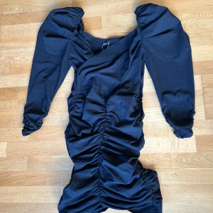 Svart långärmad klänning med puffärm från Gina Tricot - Säljer en svart långärmad klänning från Gina Tricot med puffärmar och rynkade detaljer längs hela klänningen. Klänningen har en figurnära passform och omlott-vibb upptill. Perfekt för dig som vill sticka ut lite extra!