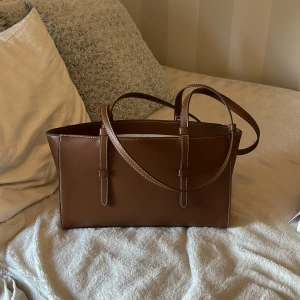 Brun axelväska i skinn - Snygg brun axelväska i skinn med stilrena vita sömmar och dubbla handtag. Väskan har en klassisk rektangulär form och passar perfekt för att bära dina vardagsprylar. Enkel och tidlös design som lyfter varje outfit. Aldrig använd 