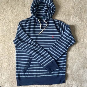 Blå- och grårandig hoodie från Polo Ralph Lauren - Säljer en blå- och grårandig hoodie från Polo Ralph Lauren med huva och dragsko. Tröjan har en broderad röd logga på bröstet och en praktisk ficka framtill. Perfekt för en avslappnad stil.
