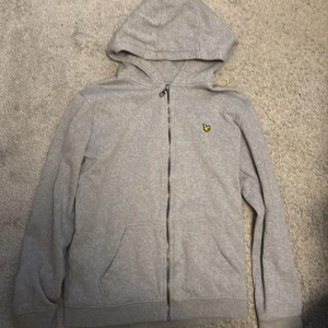 Grå hoodie från Lyle & Scott - Grå hoodie från Lyle & Scott med dragkedja och huva. Klassisk design med broderad gul logga på bröstet och två fickor framtill. Tillverkad i mjuk bomull, perfekt för chill dagar. Passar dig som gillar enkel och stilren streetwear.