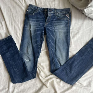 Blå Replay jeans med slitningar - Snygga blå jeans från Replay med slitningar och tvättade detaljer framtill och på bakfickorna. Klassisk femficksmodell med normal passform och raka ben. Jeansen har en mörkblå bas med ljusare partier och Replay-logga på bakfickan. Går och pruta lite 