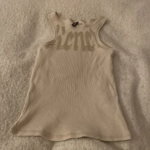 Beige ribbad linnetopp från Divided - Snygg beige ribbad linnetopp från Divided by H&M i storlek XXS. Toppen har brottarrygg och tryck framtill i en ljusare beige ton. Perfekt att styla med jeans eller kjol för en chill look.