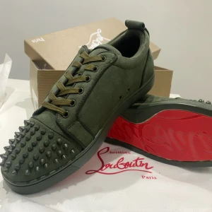 Olivgröna sneakers från Louboutin - Snygga olivgröna sneakers från Christian Louboutin i mocka med ikoniska röda sulor. Skorna har snörning och coola nitar på tån för en edgy vibe. Perfekta för dig som vill sticka ut med stilrena detaljer och exklusiv känsla.