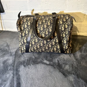 Christian Dior monogram handväska - Säljer en klassisk handväska från Christian Dior med ikoniskt monogrammönster i svart och beige. Väskan har svarta skinnhandtag och detaljer, dragkedja upptill och rymlig insida. Perfekt för dig som vill ha en statementväska med lyxig vintagekänsla.