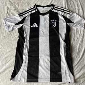 Juventus Adidas Fotbolls Tröja  - Juventus fotbollströja från Adidas i slim fit, klassiskt svartvitt randigt mönster med klubbmärke och tre stjärnor på bröstet. HELT NY OANVÄND ✅ Perfekt för dig som älskar fotboll och vill bära klubbens färger. 