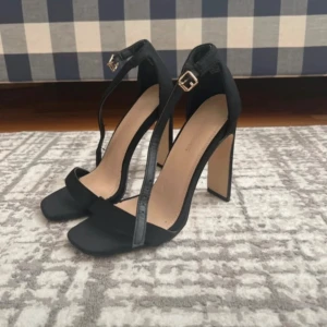 Svarta klackade sandaler med remmar - Stilrena svarta sandaler med hög klack och smala remmar över foten och runt vristen. Skorna har fyrkantig tå och justerbart spänne i guld. Perfekta för att lyfta din outfit med en elegant touch.