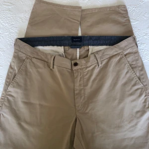 Beiga chinos från Gant - Säljer ett par klassiska beiga chinos från Gant med slim passform. Byxorna har snedställda fickor fram, bakficka med knapp och bälteshällor. Perfekta till vardags och stilrena till många outfits. Endast använda två gånger. I väldigt fint skick, nästintill nyskick. Storlek W38|L32