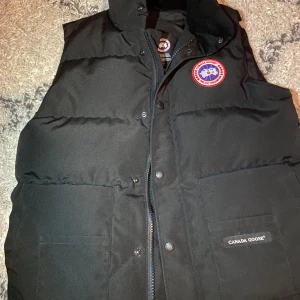 Svart dunväst från Canada Goose - Säljer en svart dunväst från Canada Goose med klassisk logga på bröstet. Västen har hög krage, två stora fickor framtill och stängs med både dragkedja och knappar. Perfekt för lager-på-lager och riktigt snygg till streetwear.