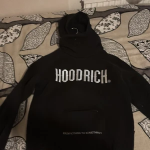 Svart HOODRICH hoodie med tryck - Svart hoodie från HOODRICH med stor vit logga framtill och texten 'FROM NOTHING TO SOMETHING' nedanför. Klassisk huva och magficka, perfekt för en streetwear-look. Skön och avslappnad passform.