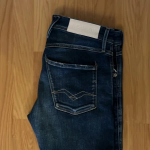 Replay Anbass jeans i mörkblå denim - Säljer ett par Replay Anbass jeans i mörkblå tvättad denim med snygga slitningar och kontrastsömmar. Klassisk femficksmodell, normal passform och raka ben. Märkespatch bak i vitt och diskret Replay-logga vid fickan. Perfekta för en avslappnad och stilren look.