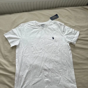 Vit t-shirt Polo Ralph Lauren M - Helt ny klassisk vit t-shirt från Polo Ralph Lauren i modellen Custom Slim Fit. T-shirten har korta ärmar, rund halsringning och den ikoniska broderade loggan i mörkblått på bröstet. Tillverkad i 100% bomull för en skön och lätt känsla.