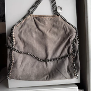 Grå axelväska Stella McCartney - Säljer en snygg grå axelväska från Stella McCartney med kedjehandtag och coola detaljer längs kanterna. Väskan är i mjuk mocka och har ett rosa innerfoder samt dragkedja på insidan. Perfekt för dig som gillar statement-accessoarer. Det finns defekter, såsom att kedjorna och tyget vid öppningen av väskan är lite missfärgade. Det är möjligt att tvätta bort missfärgning på tyget. Väskan är rätt stor 