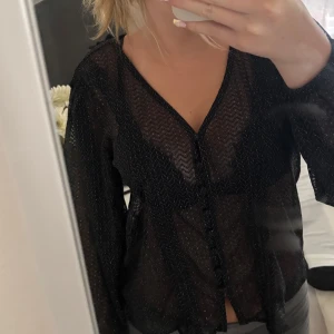 Svart glittrig blus med knappar - Säljer en transparent svart blus med glittriga detaljer och subtilt sicksackmönster. Blusen har v-ringning, långa ärmar och knappar framtill. Perfekt för dig som vill sticka ut lite extra med en snygg och trendig topp.