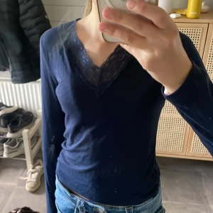 Mörkblå långärmad topp med spets - Mörkblå långärmad topp med v-ringning och snygg spetsdetalj runt halsen. Sitter figurnära och är perfekt till jeans eller kjol. Mjuk och skön i bomullsmaterial, enkel men ändå med en fin touch av spets.