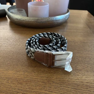 Svart och grått flätat bälte - Snyggt flätat bälte i svart, grått och vitt med detaljer i brunt skinn vid spännet. Bältet har ett coolt randigt mönster och ett silvrigt spänne. Perfekt accessoar för att lyfta din outfit med en modern touch.