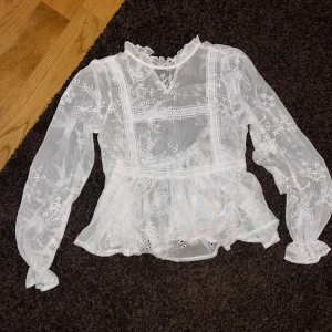 Vit spetsblus från NA-KD - Superfin vit blus från NA-KD i transparent spets med broderade blommor och volangdetaljer. Hög krage och långa ärmar med romantisk känsla. Perfekt att styla med jeans eller kjol för en trendig look.