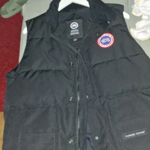 Svart dunväst från Canada Goose - Säljer en svart dunväst från Canada Goose med klassisk logotyp på bröstet och diskret varumärkesmärke vid fickan. Västen har knappar och dragkedja framtill samt två stora fickor.