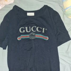 Svart t-shirt från Gucci - Svart t-shirt från Gucci med klassisk logga och färgade ränder i grönt och rött över bröstet. T-shirten har rund halsringning och korta ärmar. Perfekt för dig som gillar exklusiva märken och stilren design.