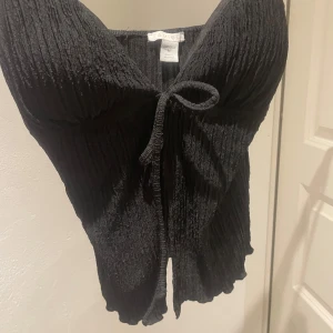 Svart ribbad linnetopp med knyt - Säljer en svart ribbad linnetopp med smala axelband och knyt framtill. Den är även öpped i magen. Har också som ”inlägg” vid brösten vilket gör att man ej behöva ha bh. Hör av er om ni vill ha flera bilder! Köpte i Tyskland så varumärket är oklart.