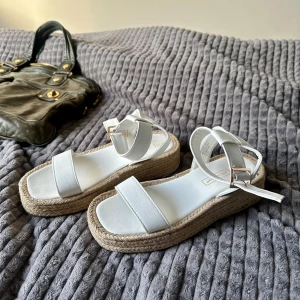 Vita sandaler - Säljer ett par vita sandaler med breda remmar och justerbart spänne vid ankeln. Skorna har en snygg platåsula i flätad jute som ger en somrig känsla. Perfekta till varma dagar och lätta att matcha med olika outfits. Aldrig använda💕