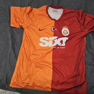 Galatasaray fotbollströja från Nike - Galatasaray matchtröja från Nike med klassisk halvorange och röd design, korta ärmar och Dri-Fit material. Klubbmärke och sponsortryck på bröstet, samt tryck på ryggen. Perfekt för fans av laget.