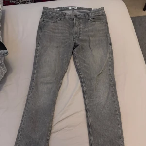 Grå jeansbyxor från Jack & Jones - Säljer ett par grå jeansbyxor från Jack & Jones med klassisk femficksdesign och raka ben. Byxorna har normal passform och stängs med dragkedja och knapp. Perfekta till vardags för en stilren look. Storlek L30/W28